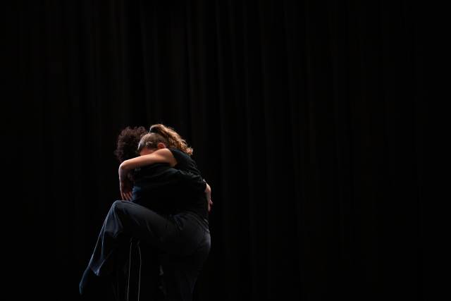 two women wearing dark colored clothing embrace with one dance legs wrapped around the other's body