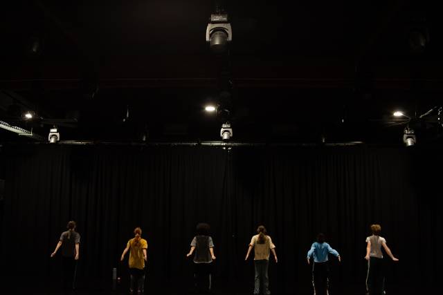 6 dancers face away from the camera in a straight line spread across the upstage in front of a black curtain.  Their arms swing back behind them.