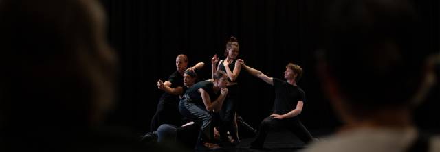 6 dancers wearing all black in different poses while connected to each other by one limb. two people facing away from the camera observe