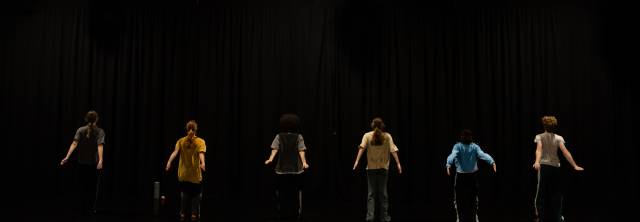 6 dancers face away from the camera in a straight line spread across the upstage in front of a black curtain.  Their arms swing back behind them.