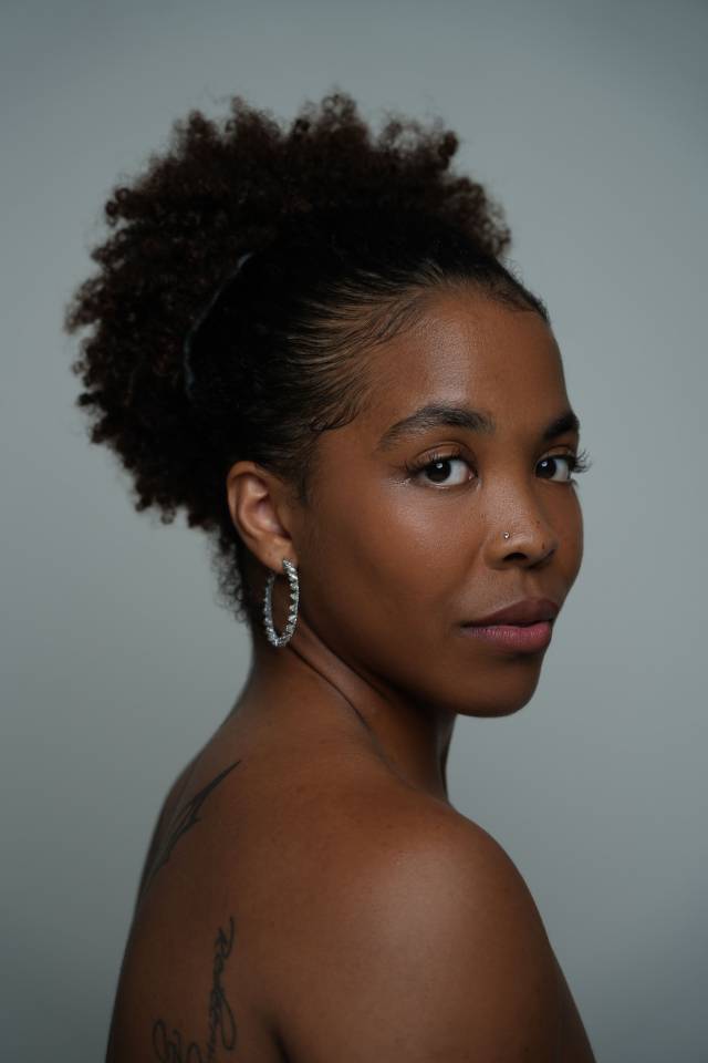 three-quarter closeup profile of a brown skinned woman with her hair pulled back in a high curly ponytail. bare shouldered and wearing a silver hoop earring and a small silver nose ring