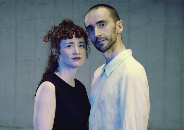 two people look towards the camera. one olive-skinned person has curly red hair and wears a sleeveless black top. the other is olive-skinned with a black mustache and beard and wears a white button down shirt