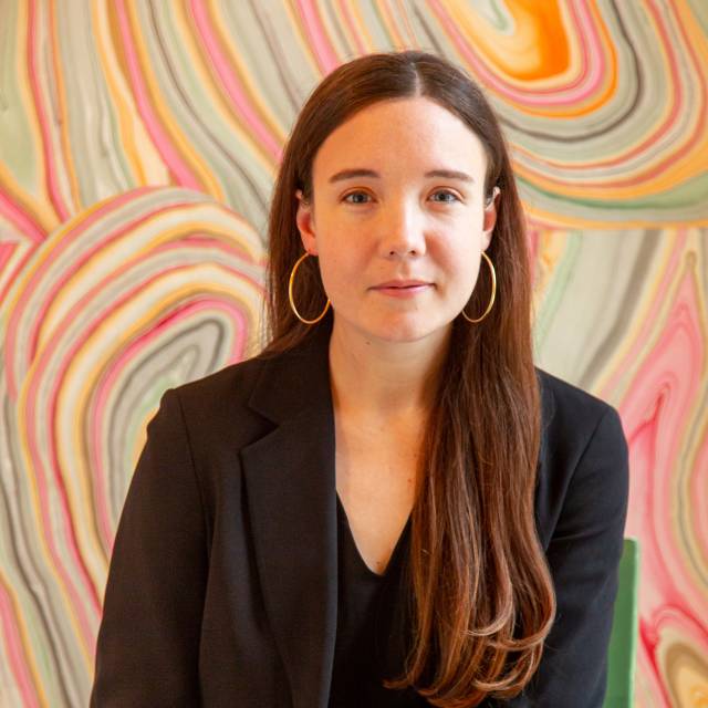 fair-skinned woman woman with long brown hair, gold earrings, black shirt and black blazer poses in front of a colorful background with wavy lines