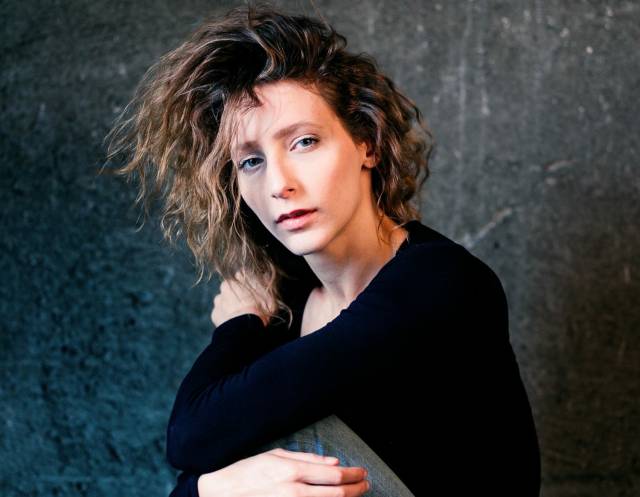 Woman with wavy brownish hair and wearing a black long sleeve shirt looks into camera with her arms loosely wrapped around her