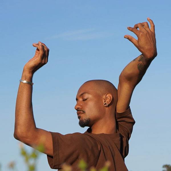 Brown skinned man with closely shaved head contorts his arms toward the sky out in nature