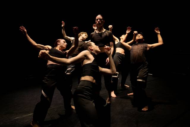 a person looks out into the distance while being surrounded by a group of five bodies with arms flailed backwards