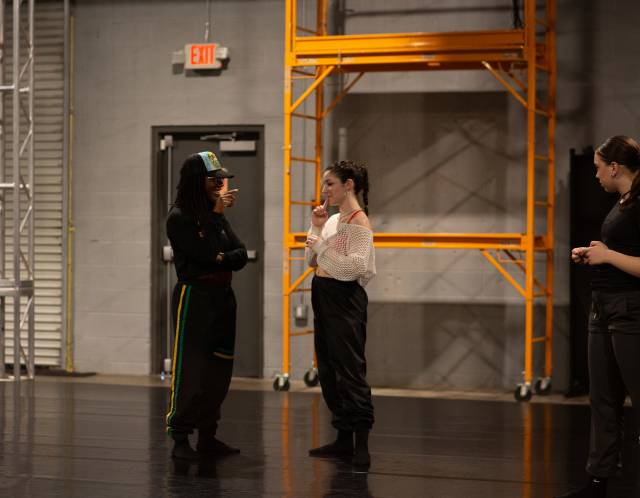 brown skinned woman wearing all black with a baseball cap speaks to an olive skinned student with a braided high ponytail