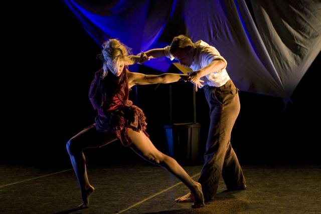 fair-skinned woman with short red bustled dress in a lunge, her arms pulled back by fair-skinned main in white button-down and brown pants