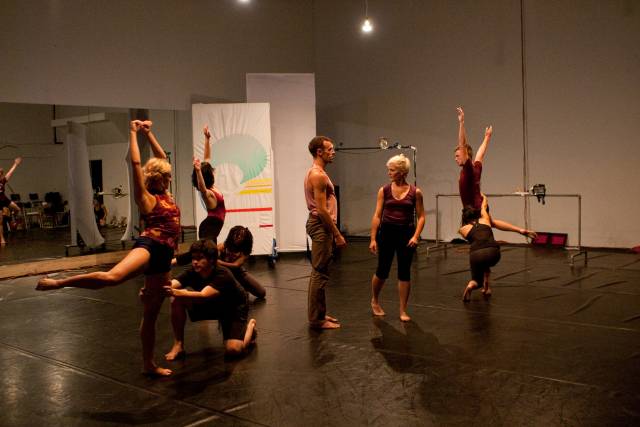 a man and woman stand in the center of 3 couples. one person in each coupling is in arabesque with their arms in the air.  the other person in the coupling holds their partner's ankle.
