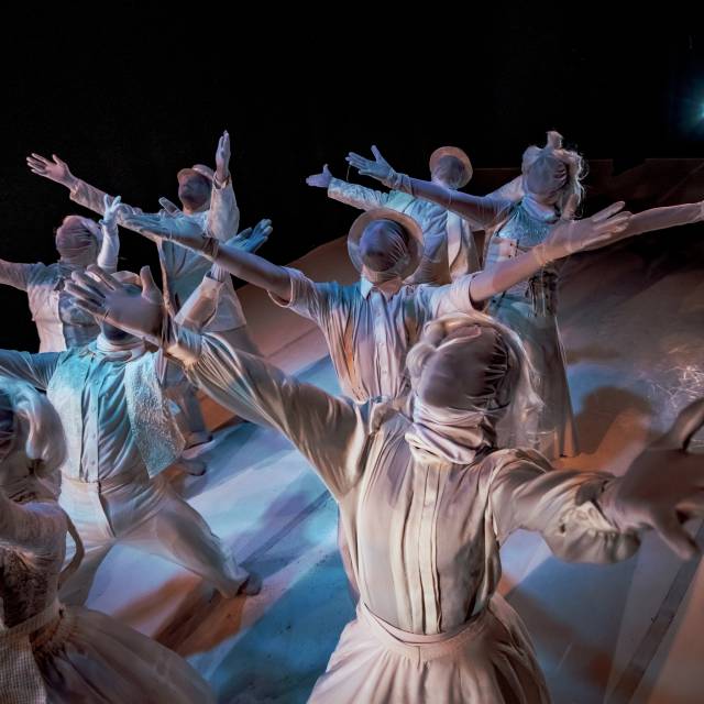8 dancers dressed in all white clothing with white wigs and hats and white mesh covering their faces and white gloves covering their hands.  standing on a white stage with their arms outstretched.