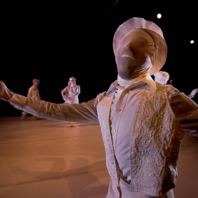 dark brown person dressed in white clothing from head to toe, including a white hat and white mesh over their face. arms outstretched with a cheshire cat grin.