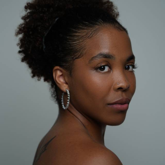three-quarter closeup profile of a brown skinned woman with her hair pulled back in a high curly ponytail. bare shouldered and wearing a silver hoop earring and a small silver nose ring