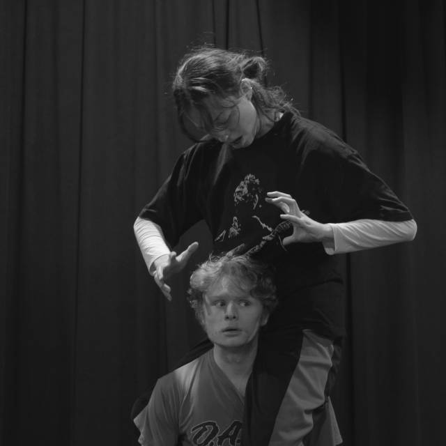 A woman straddles the shoulder of a man and cowers over him with hands hovering around his head