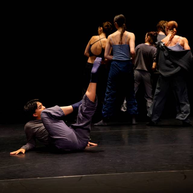 a man on the floor stretches one leg to the sky while a group of four people stand upstage turned away from him