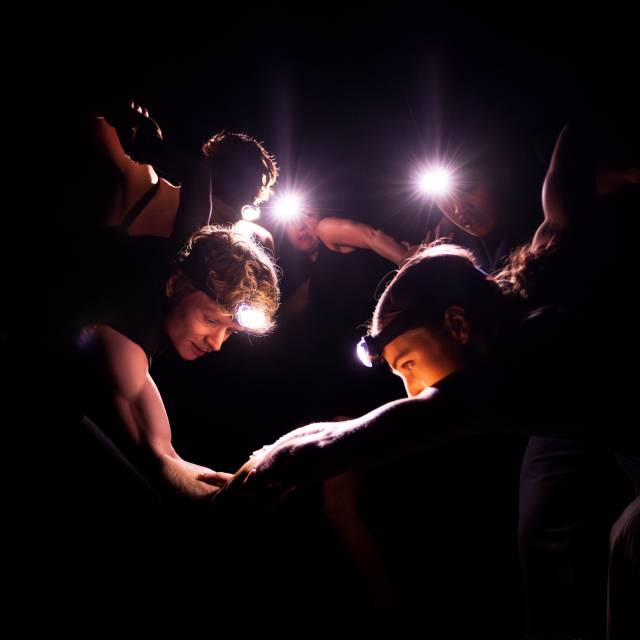 4 dancers link arms in a dark environment with headlamps on
