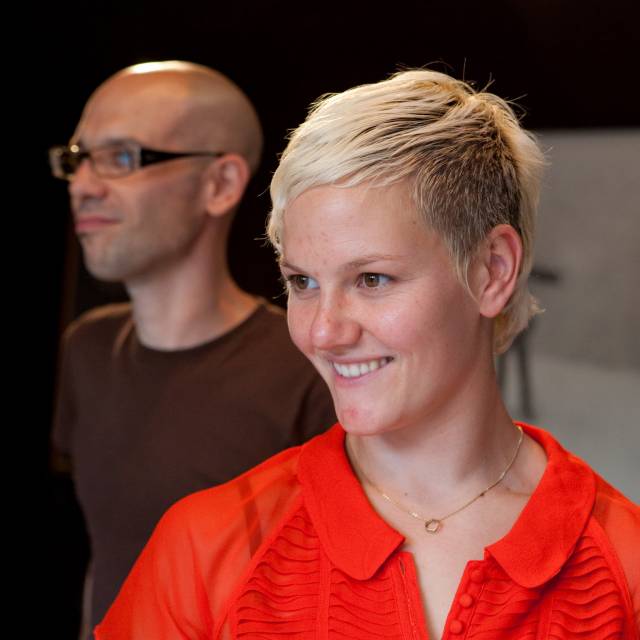a olive skinned bald man wearing black glasses and a brown shirt standing diagonally behind a fair skinned smiling woman with short blonde hair and a orange collared shirt
