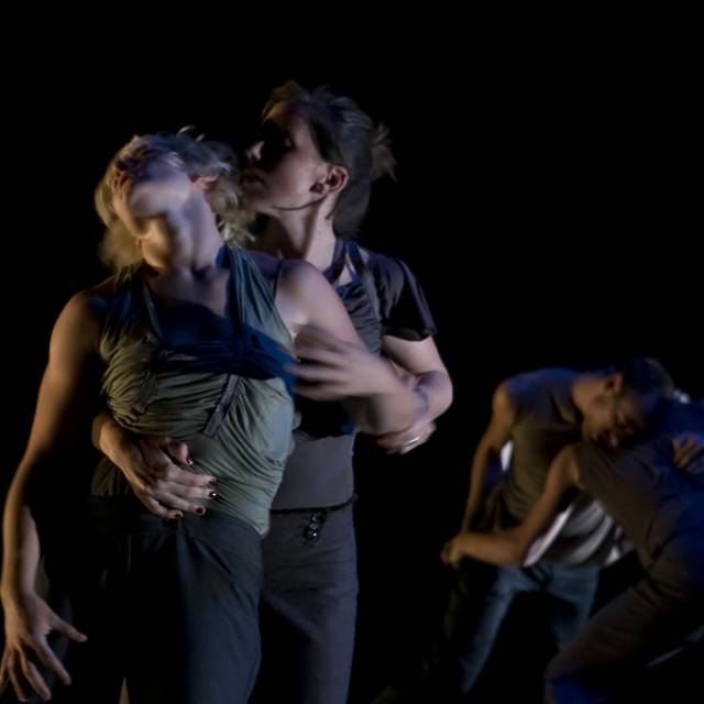 two fair skinned women in grey. one hugs the other from behind.