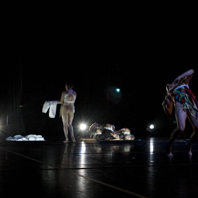 a figure wearing colorful pieces of material hinges while another figure walks away carrying a white piece of clothing