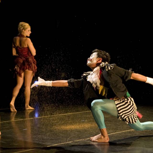 medium-skinned guy dressed in a brightly colored jester costume with a painted face throws dust while in a lunge