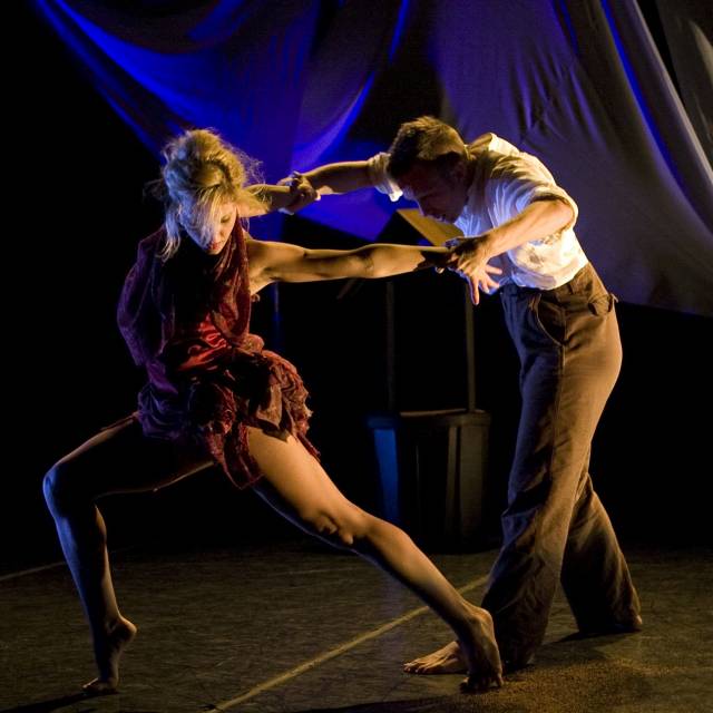 fair-skinned woman with short red bustled dress in a lunge, her arms pulled back by fair-skinned main in white button-down and brown pants