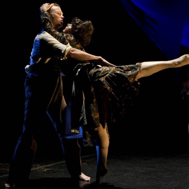 fair-skinned woman in floral dress in an arabesque while grabbing onto the head of a fair-skinned main in a white button down and brown pants
