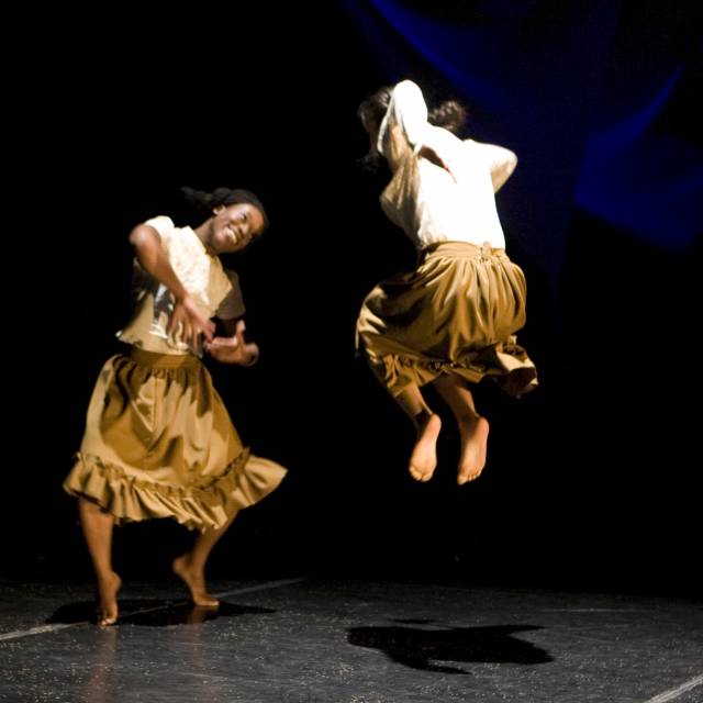 brown skinned woman and medium skinned woman smile and face off while jumping up and down. they wear a white top and brown skirt.