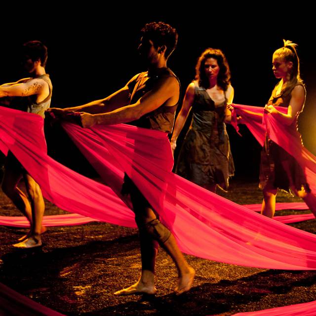 3 people stand in black sand draped in red cloth while a woman walks amongst them
