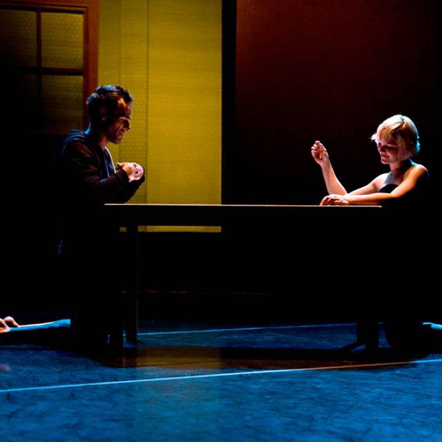 a fair skinned man and woman kneel across from each other at separate ends of a wooden table