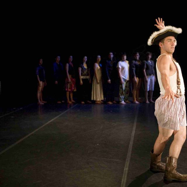 fair skinned man in boxers, a pirate hat, and cowboy boots strikes a pose while a group of people watch in the background