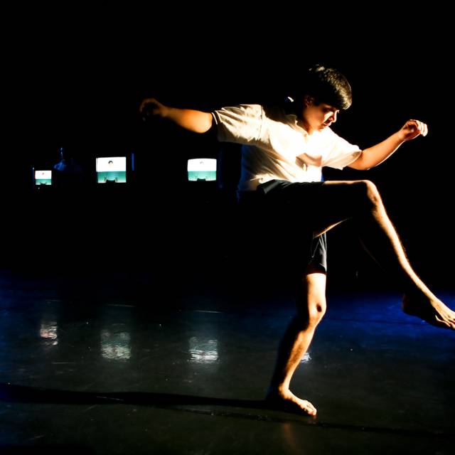 man balances on one leg with several tvs behind him