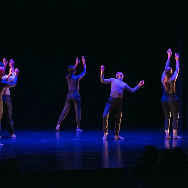 four dancers face upstage with arms raised while one dancer faces downstage with arms raised