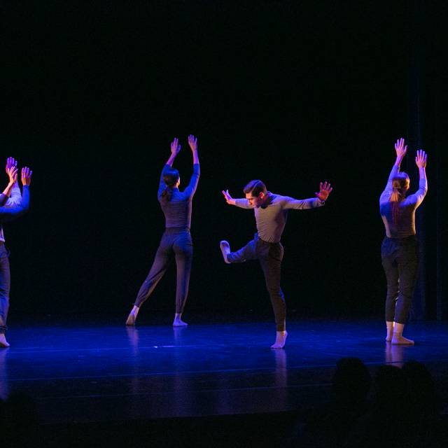 a man balances on one leg while four dancers face upstage with arms raised