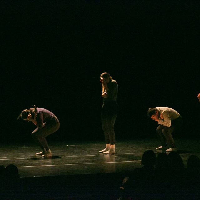 four dancers crouch down covering their faces while one dancer in the center stands while covering her face