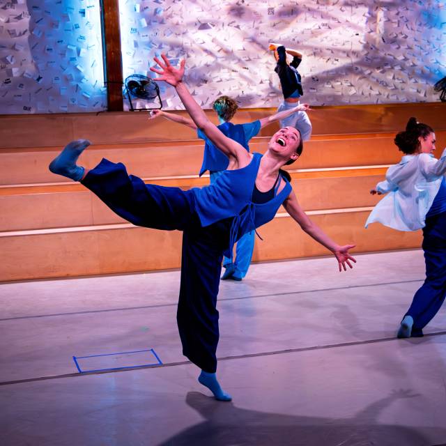 a woman dressed in blue happily stands on one leg with arms outstretched. others in mid groove behind her.