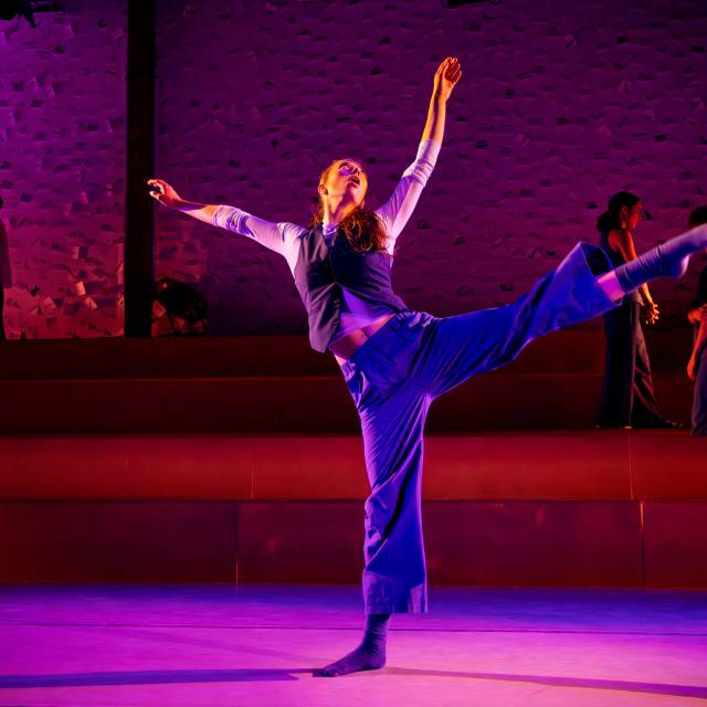a woman dressed in blue does an arabesque with outstretched arms